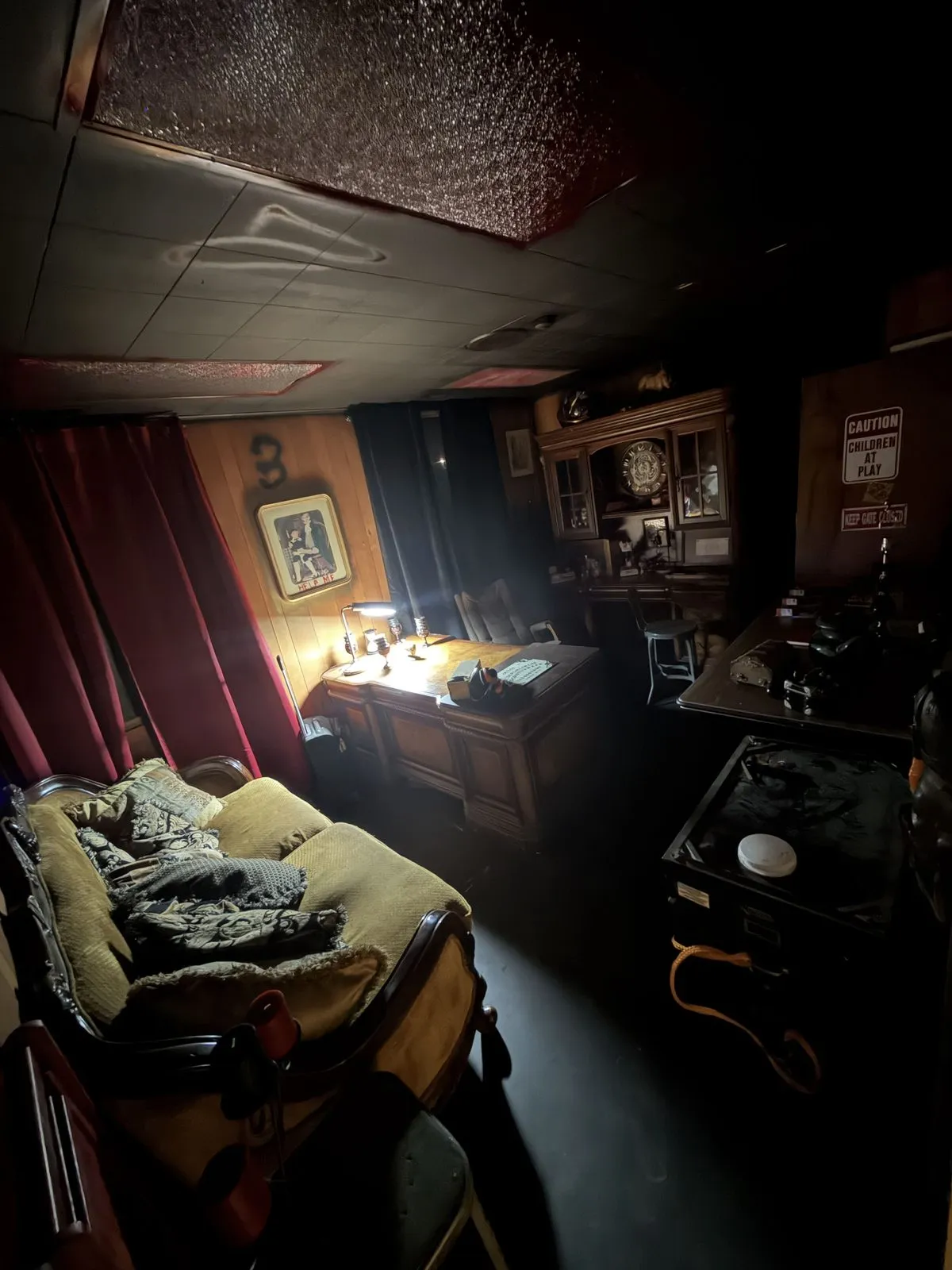 A study lit by a single desk lamp: a wooden desk, a velvet chaise with a blanket, and red curtains